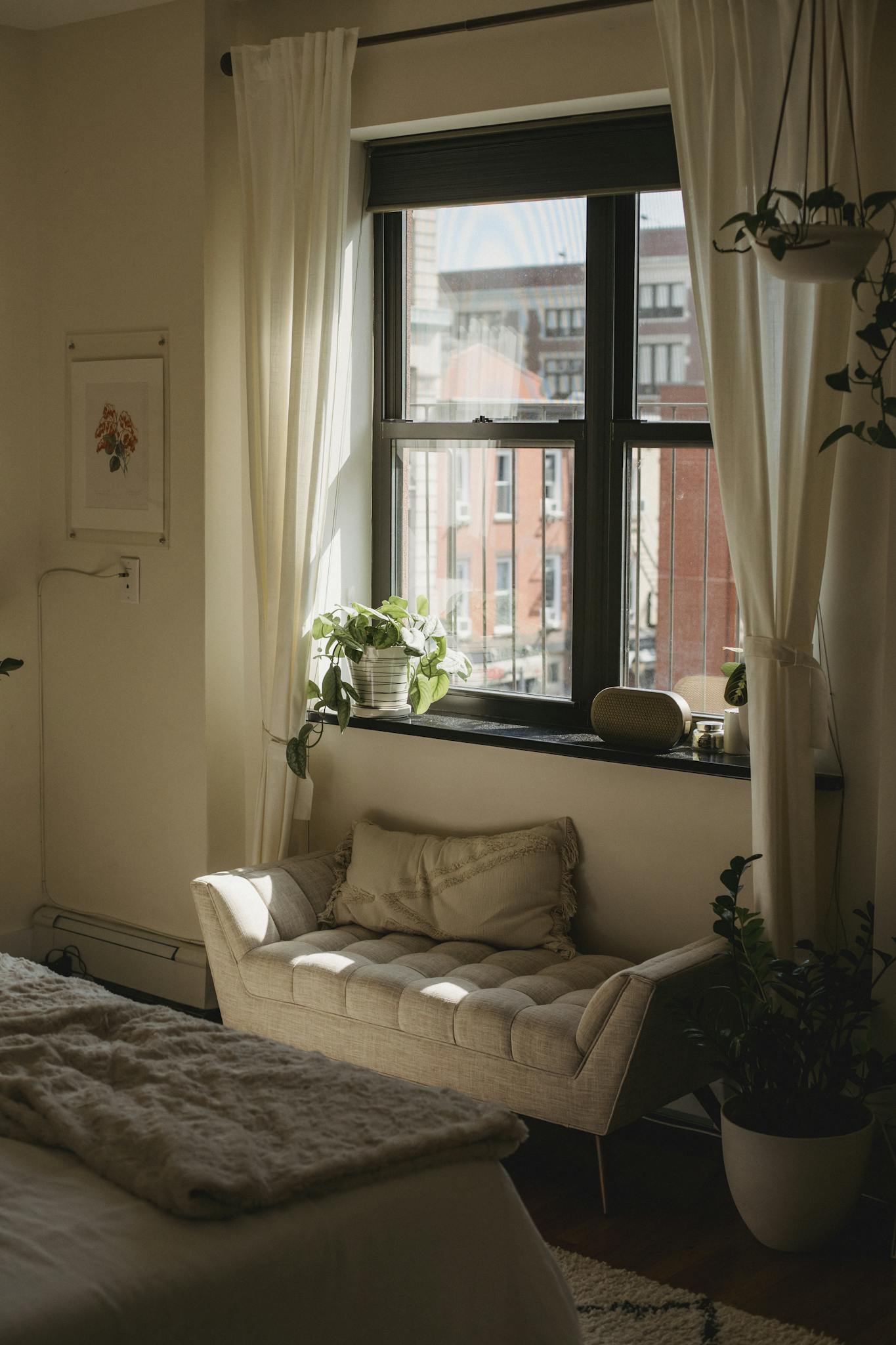 Une chambre confortable avec un éclairage tamisé dispose d'un canapé beige sous une fenêtre. Les fenêtres drapées de rideaux laissent entrer la lumière naturelle et une plante en pot ajoute de la verdure. Un couvre-lit moelleux et une vue sur la ville complètent cette scène apaisante.