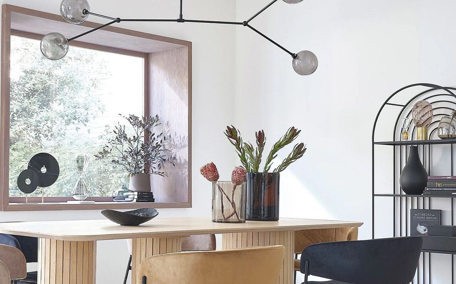 Une salle à manger moderne avec une table en bois et des chaises mixtes. Des vases décoratifs avec des plantes sont posés sur la table. Un lustre géométrique est suspendu au-dessus. Une bibliothèque avec des objets de décoration se trouve à droite et une grande fenêtre à gauche laisse entrer la lumière naturelle.