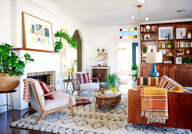 Un salon dynamique au style moderne du milieu du siècle. Il comprend un tapis douillet, des meubles en bois, des coussins colorés et des œuvres d'art abstraites. Les plantes ajoutent de la verdure et un lustre est suspendu au plafond. Un mélange de tons chauds et neutres crée une ambiance accueillante.